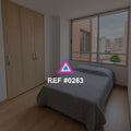 Ventana al Barrio. Una cama gris junto a un armario de madera y una ventana hacia edificios de ladrillo.