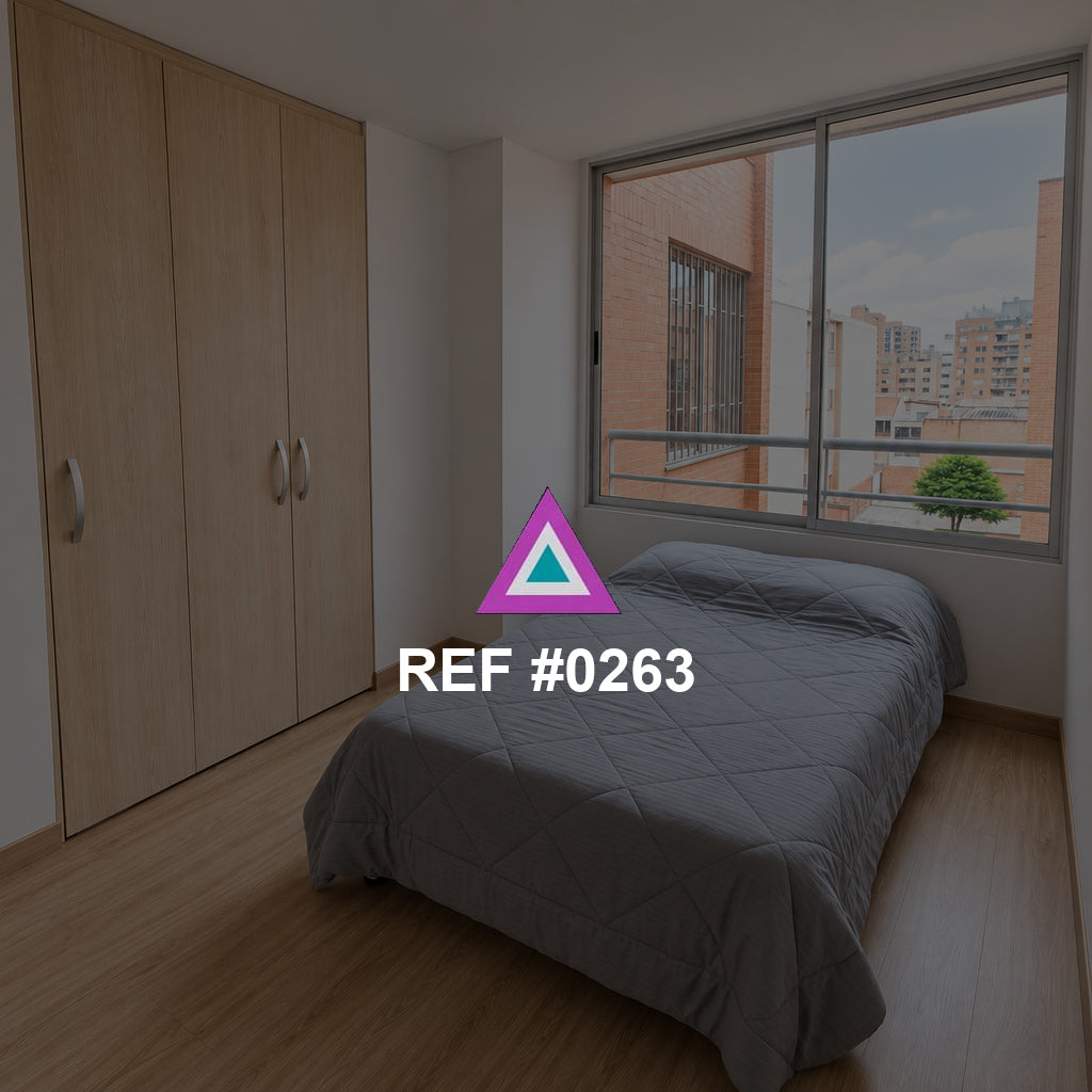 Ventana al Barrio. Una cama gris junto a un armario de madera y una ventana hacia edificios de ladrillo.
