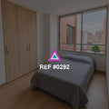 Ventana al Barrio. Una cama gris junto a un armario de madera y una ventana hacia edificios de ladrillo.